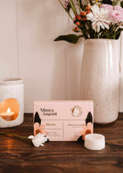 Sur une table en bois, entourée de fleurs, d'une petite bougie blanche et d'un vase de fleurs variées, repose une boîte de fondants de cire de soya Bloom de Mimi & August. Ces cubes écoresponsables offrent un parfum longue durée pour toute pièce.