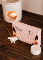 Une boîte « Mimi & August Bloom » repose sur une surface en bois avec une fleur blanche, tandis qu'un fondant de cire de soya écoresponsable et un bougeoir avec une bougie allumée offrent un parfum longue durée en arrière-plan.