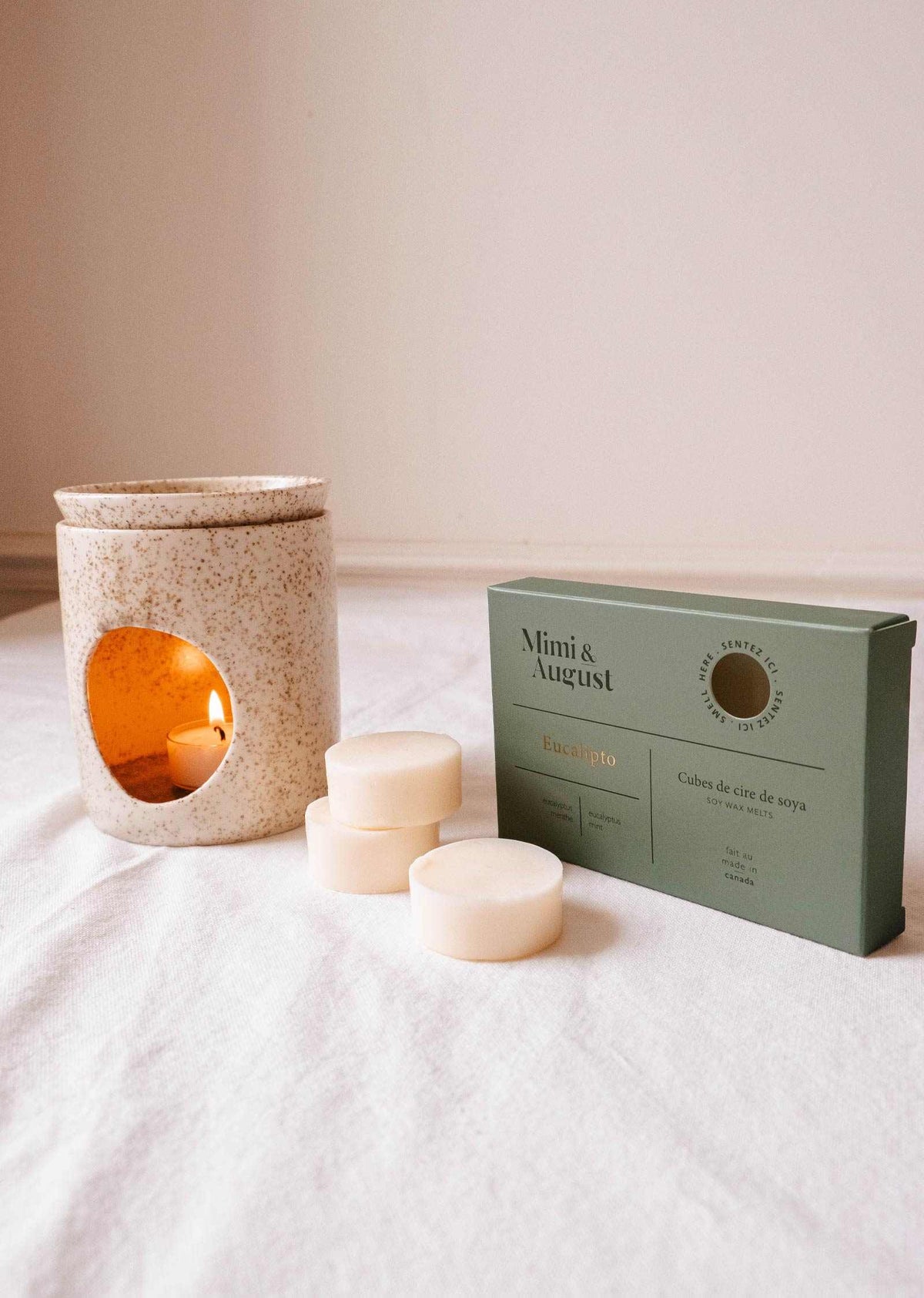A speckled ceramic candle warmer with a lit candle is positioned beside a box marked "Soy Wax Melts - Eucalipto" by Mimi & August. Three eucalyptus soy wax melts are arranged in front of the box on a white surface, providing a natural fragrance that soothes the senses.