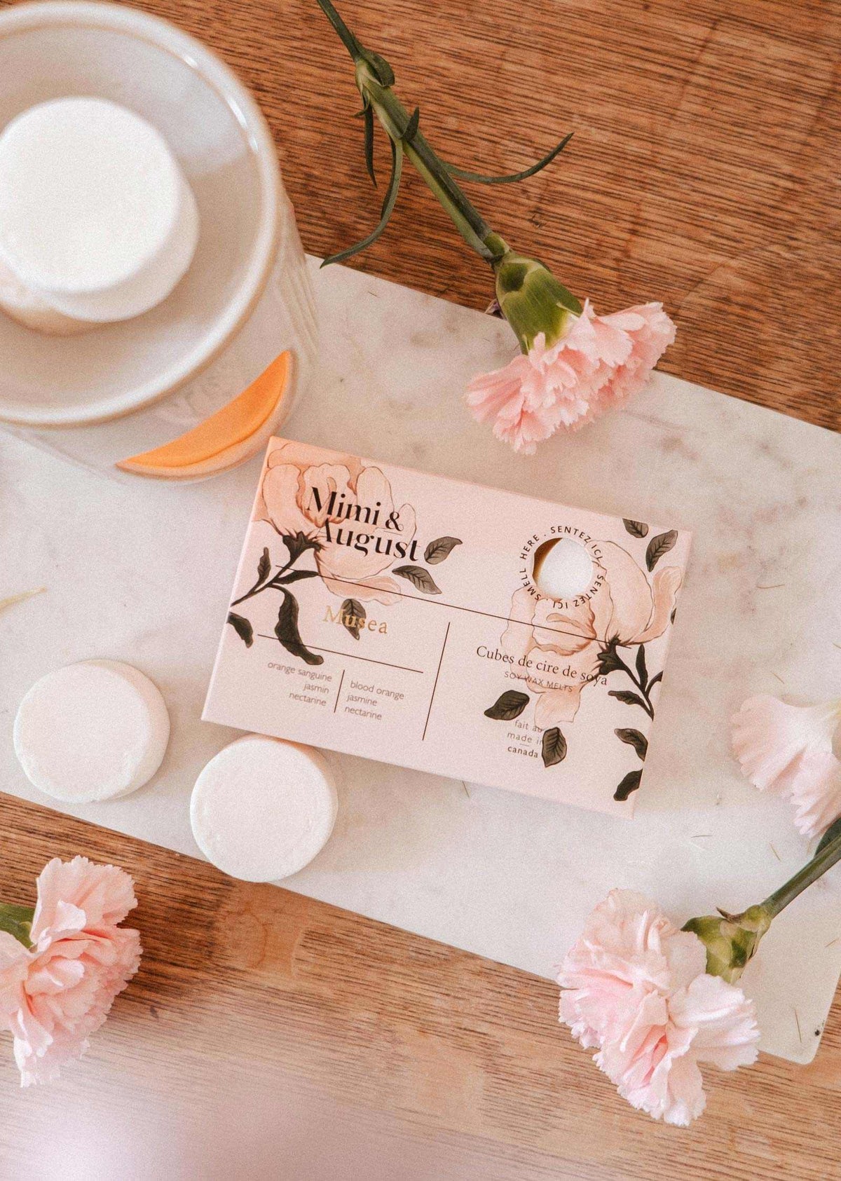 Une boîte de fondants de cire de soya Musea de Mimi & August à la fragrance non toxique est présentée sur une planche en marbre à côté d'œillets roses, d'un pot et de trois fondants de cire sur une surface en bois.