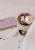 A beige wax melt burner with eco-friendly Mimi & August Soy Wax Melts - Lilac and dried flowers sits by a matching lilac box, filling the air with long-lasting fragrance atop a white fabric surface.