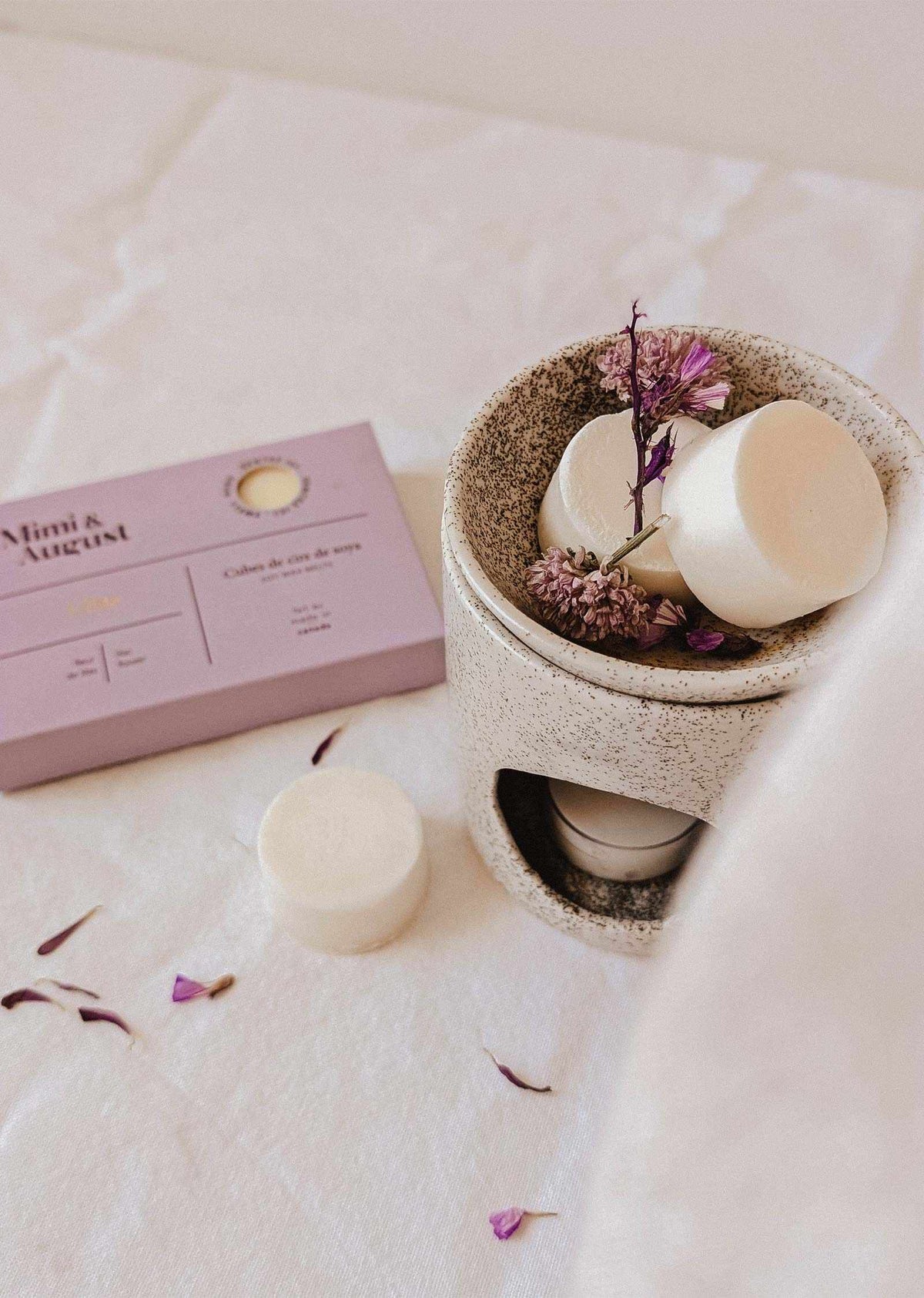 A beige wax melt burner with eco-friendly Mimi & August Soy Wax Melts - Lilac and dried flowers sits by a matching lilac box, filling the air with long-lasting fragrance atop a white fabric surface.