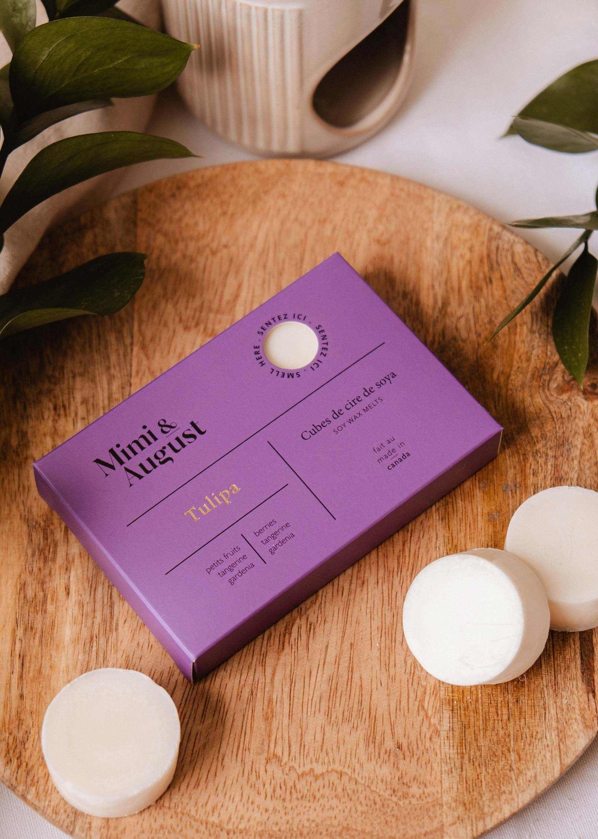 A box of Mimi & August Soy Wax Melts - Tulipa in purple rests on a round wooden tray with three white natural soy wax melts and green leaves nearby.