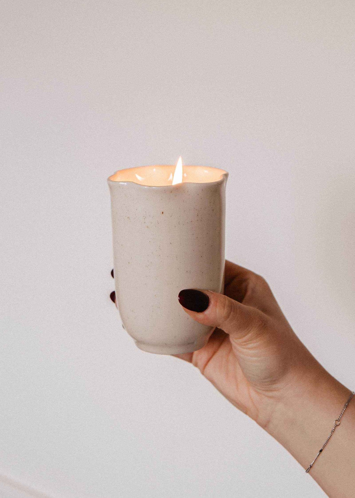 Bougie Orange Épicée dans une tasse en céramique crème mouchetée, sans anse, au rebord évasé en pétales et mèche allumée.