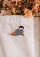 The Sir Moustache Enamel Pin by Mimi & August is a whimsical accessory for animal lovers, featuring a monkey with a white mustache on white fabric, set against dried flowers.