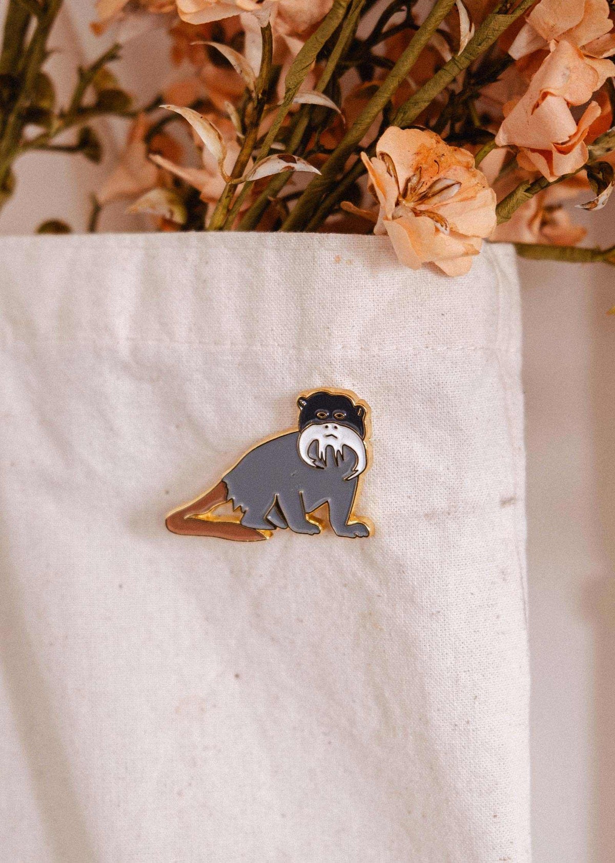 Épinglette Tamarin de Mimi & August est un accessoire plein de fantaisie pour les amoureux des animaux. Elle met en vedette un petit singe au joli moustache blanc, posé sur un fond de tissu blanc, accompagné de fleurs séchées