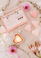 The Mimi & August "Little Hearts – Tealight Candle Set" features heart-shaped soy candles, a lit candle in a heart dish, pink flowers, and white petals elegantly displayed for romantic home decor.