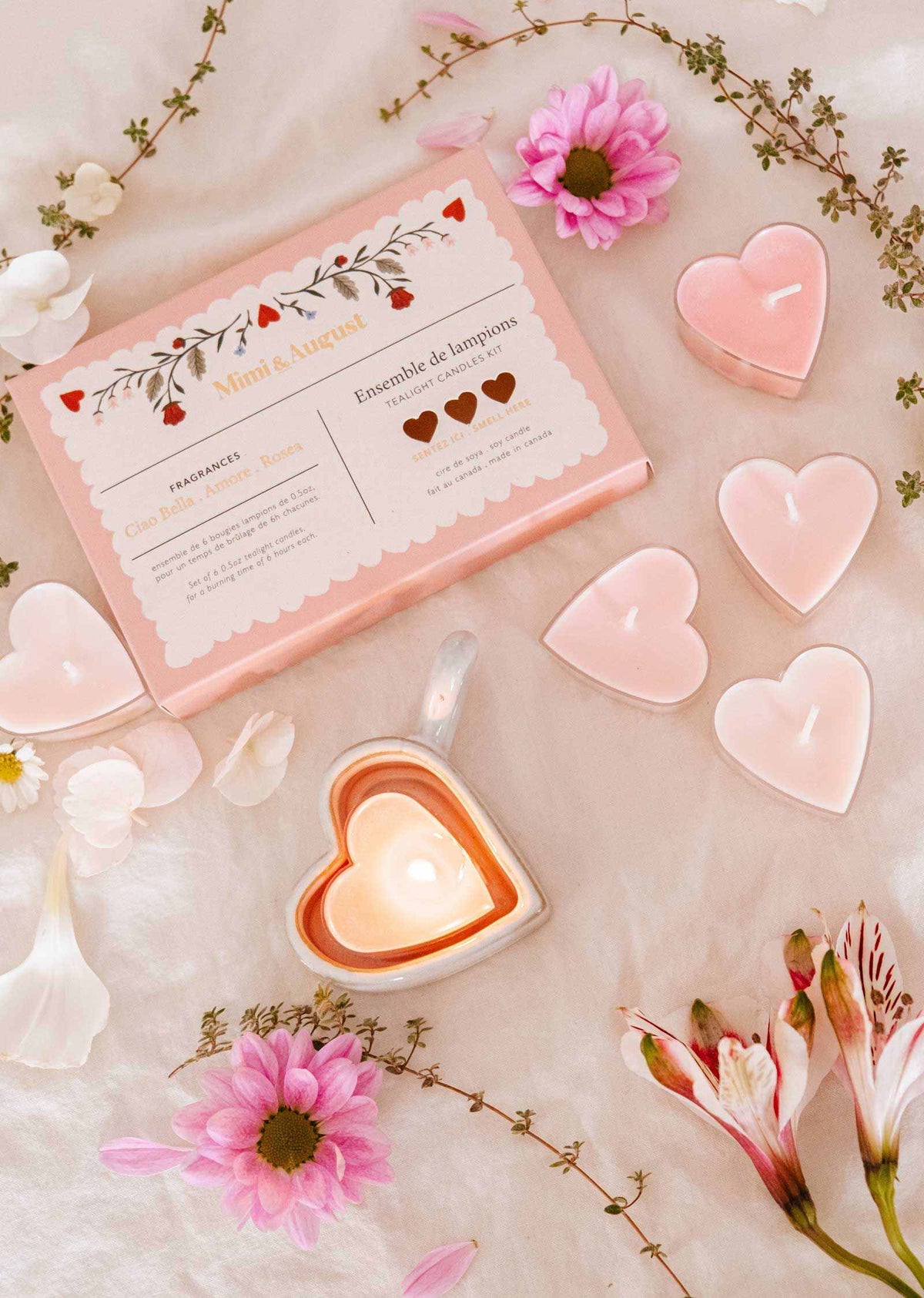 The Mimi & August "Little Hearts – Tealight Candle Set" features heart-shaped soy candles, a lit candle in a heart dish, pink flowers, and white petals elegantly displayed for romantic home decor.