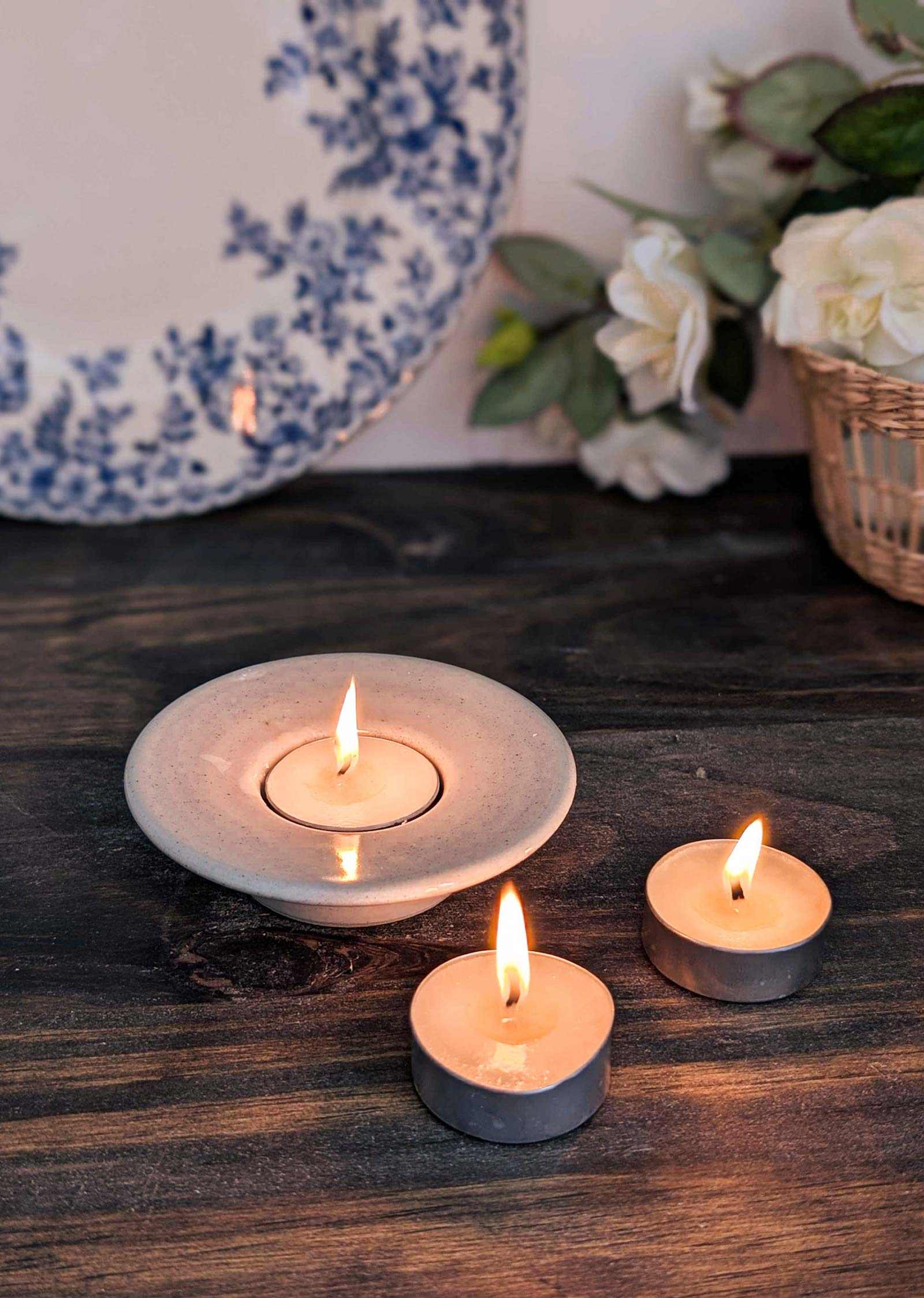Three tea lights from the Mimi & August Spring Discovery Kit glow on a wooden surface, with a floral plate and a basket of white flowers in the background—perfect for enjoying fresh spring scents.