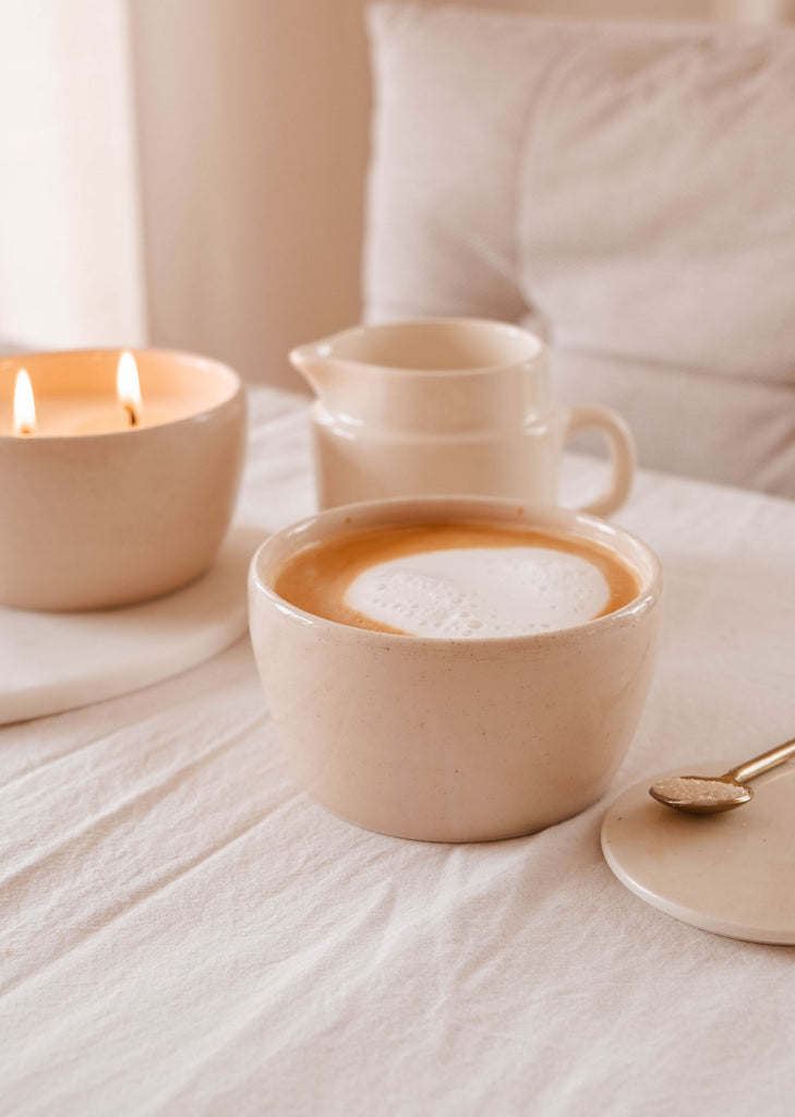 A ceramic cup of coffee with froth on a tablecloth, accompanied by a Mimi & August Bloom Candle and a creamer pitcher, evoking a cozy atmosphere.