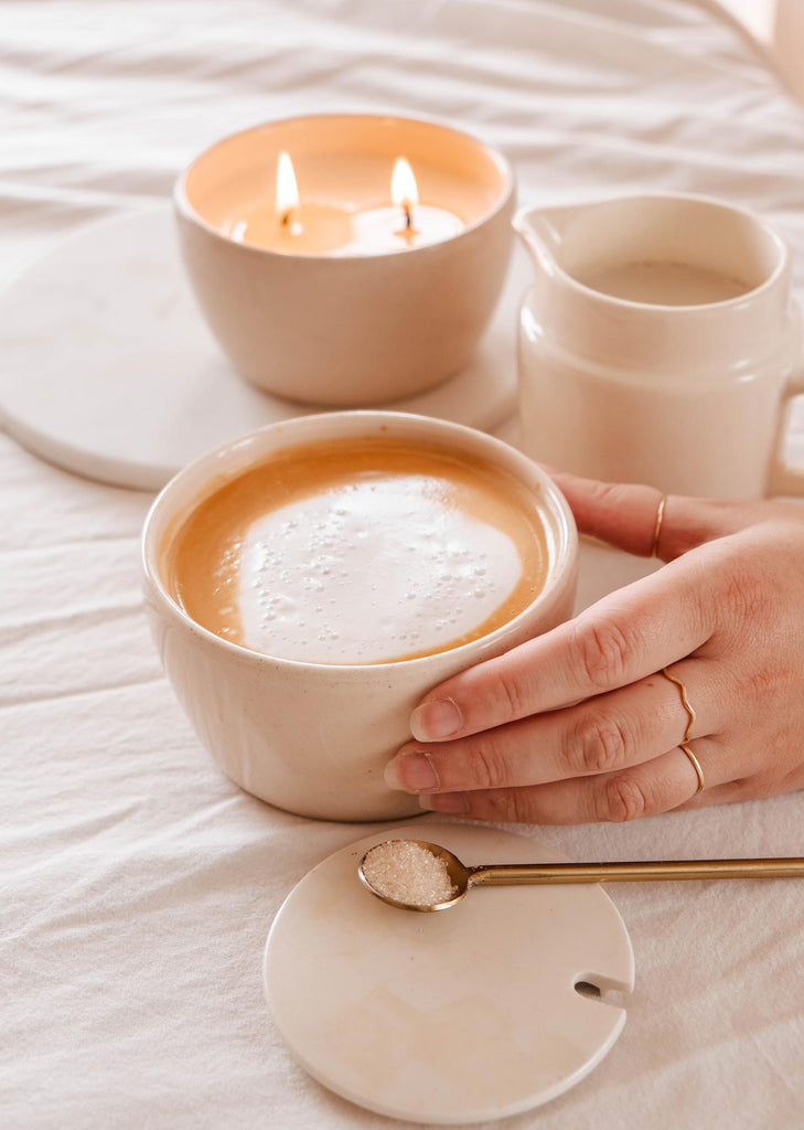A person's hands holding a ceramic mug of coffee, with a Mimi & August Bloom Candle, another mug, and a saucer with a spoon on a bed with white linen.