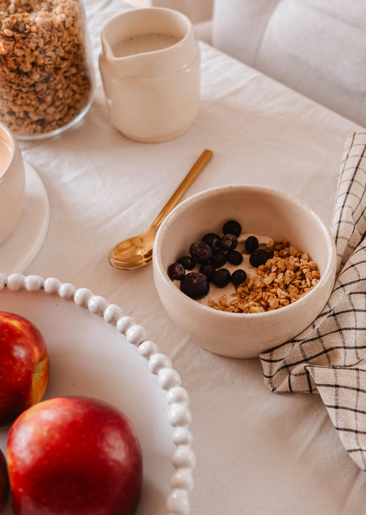 Un bol de granola avec des myrtilles, accompagné de pommes, de la bougie Bloom de Mimi & August, d'une tasse et d'une cuillère sur une nappe soigneusement disposée et ornée d'art fonctionnel.