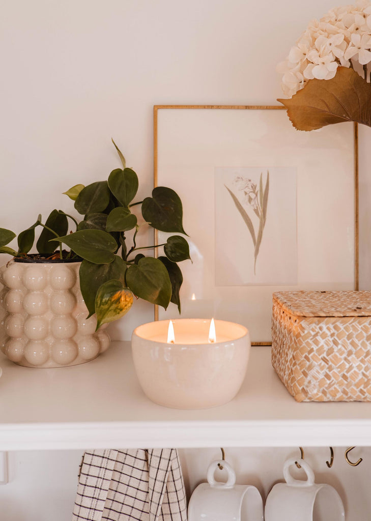 A cozy home shelf display with a Mimi & August Bloom Candle, potted plants, framed artwork, and a wicker box, conveying a warm, serene ambiance.