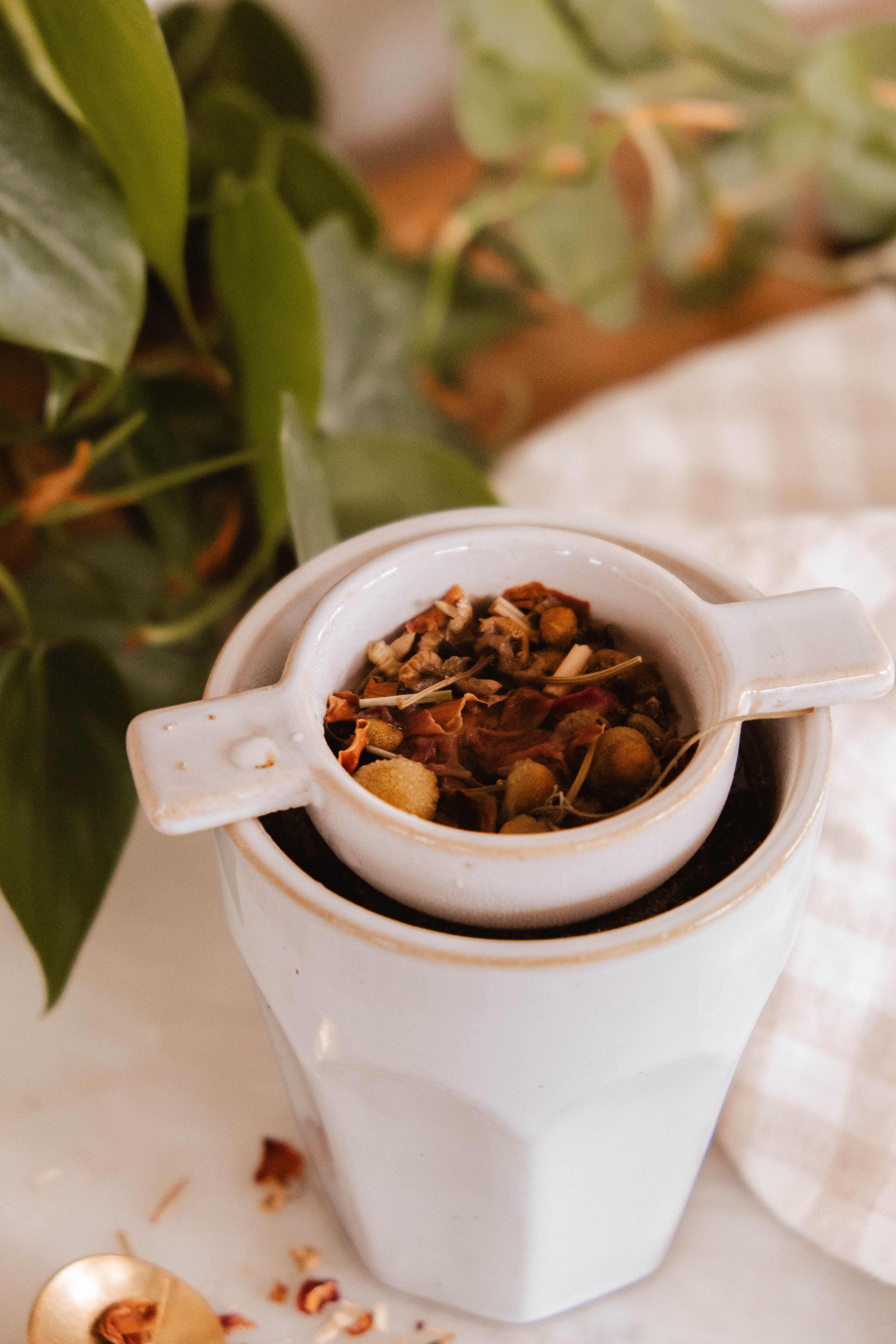 The Infusion Nest – Ceramic Tea Strainer by Mimi & August, filled with loose leaves, rests atop a white mug. A green plant and a checkered cloth add to the scene.