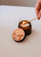 A hand lights the Mimi & August Topaze - 2 oz Soy Candle in a brown glass jar on a beige surface, filling your space with sandalwood and vanilla fragrance.
