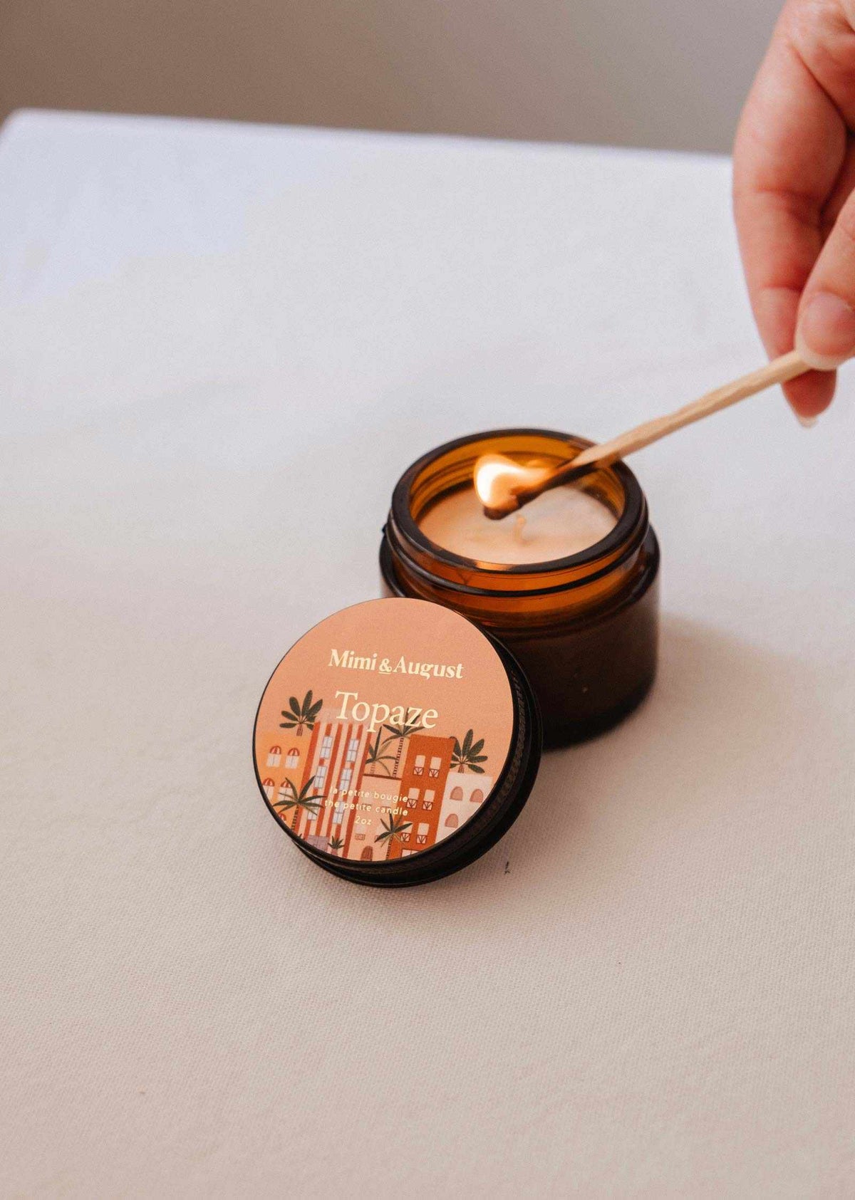 A hand lights the Mimi & August Topaze - 2 oz Soy Candle in a brown glass jar on a beige surface, filling your space with sandalwood and vanilla fragrance.