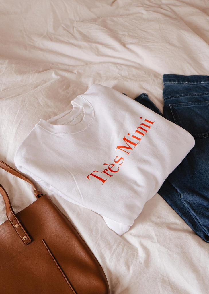 A comfortable white Très Mimi Sweatshirt and jeans, laid neatly on a bed, with a stylish Mimi & August handbag.
