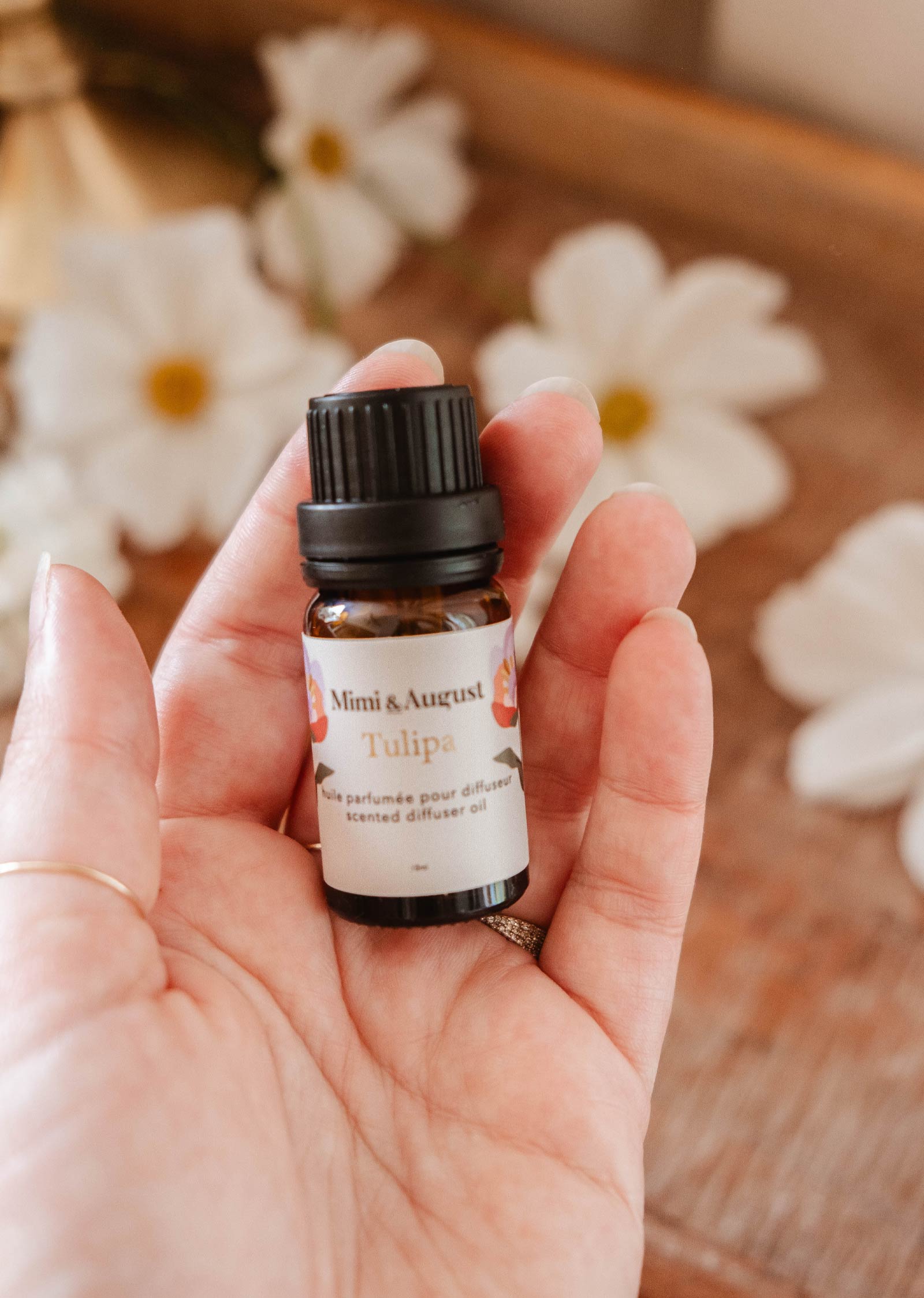 Hand holding a small brown bottle labeled "Mimi & August Scented Oil - Tulipa" amid a background with white flowers and light floral notes.