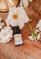 A small bottle of Scented Oil - Tulipa labeled "Mimi & August," exuding light floral notes, is surrounded by white flowers on a wooden surface.