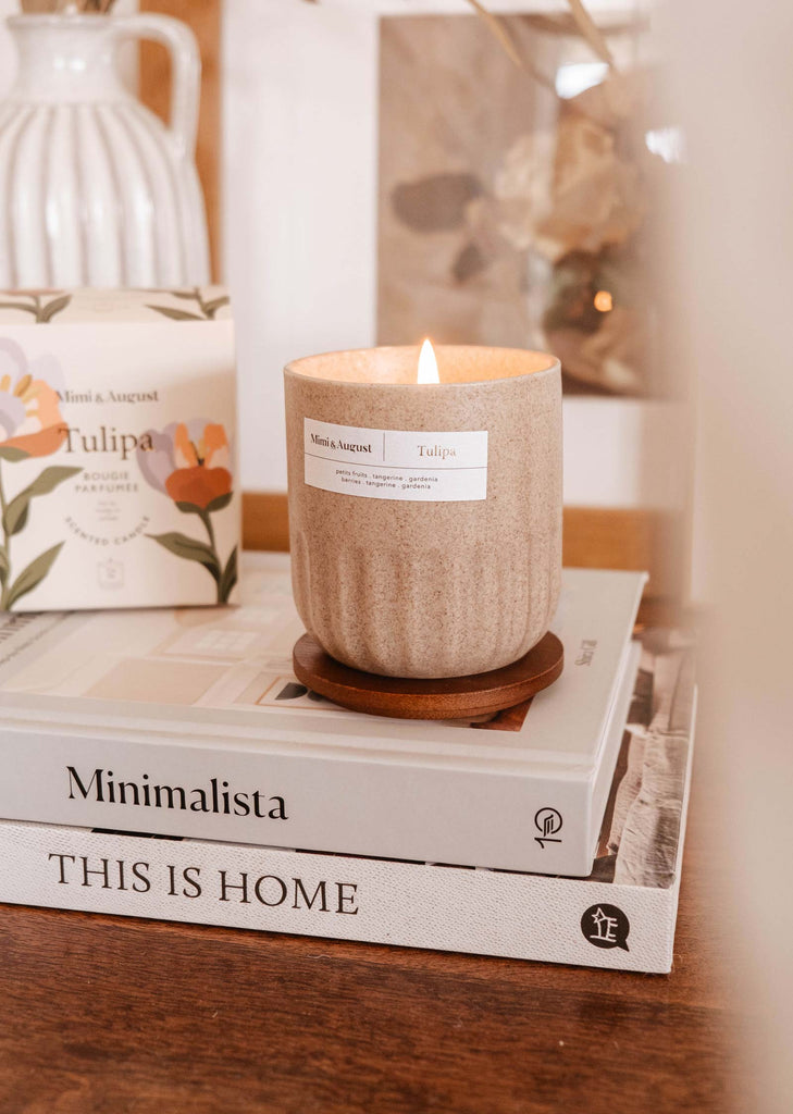 Une bougie parfumes Tulipa de Mimi & August sur un petit support en bois, placée sur une pile de livres portant les titres "minimalista" et "this is home" sur une table confortable.