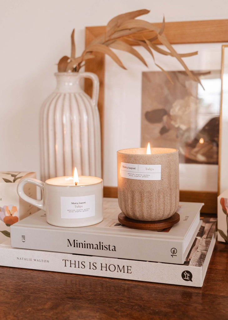 Bougies fragrance tulipa sur un livre intitulé "minimalista : this is home" avec un vase en bois, un cadre photo et une tasse à proximité sur une table. (Marque : Mimi & August)