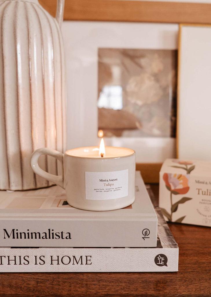 A Mimi & August Tulipa reusable candle on top of a stack of books titled "minimalista" and "this is home," next to a framed photo and a vase.