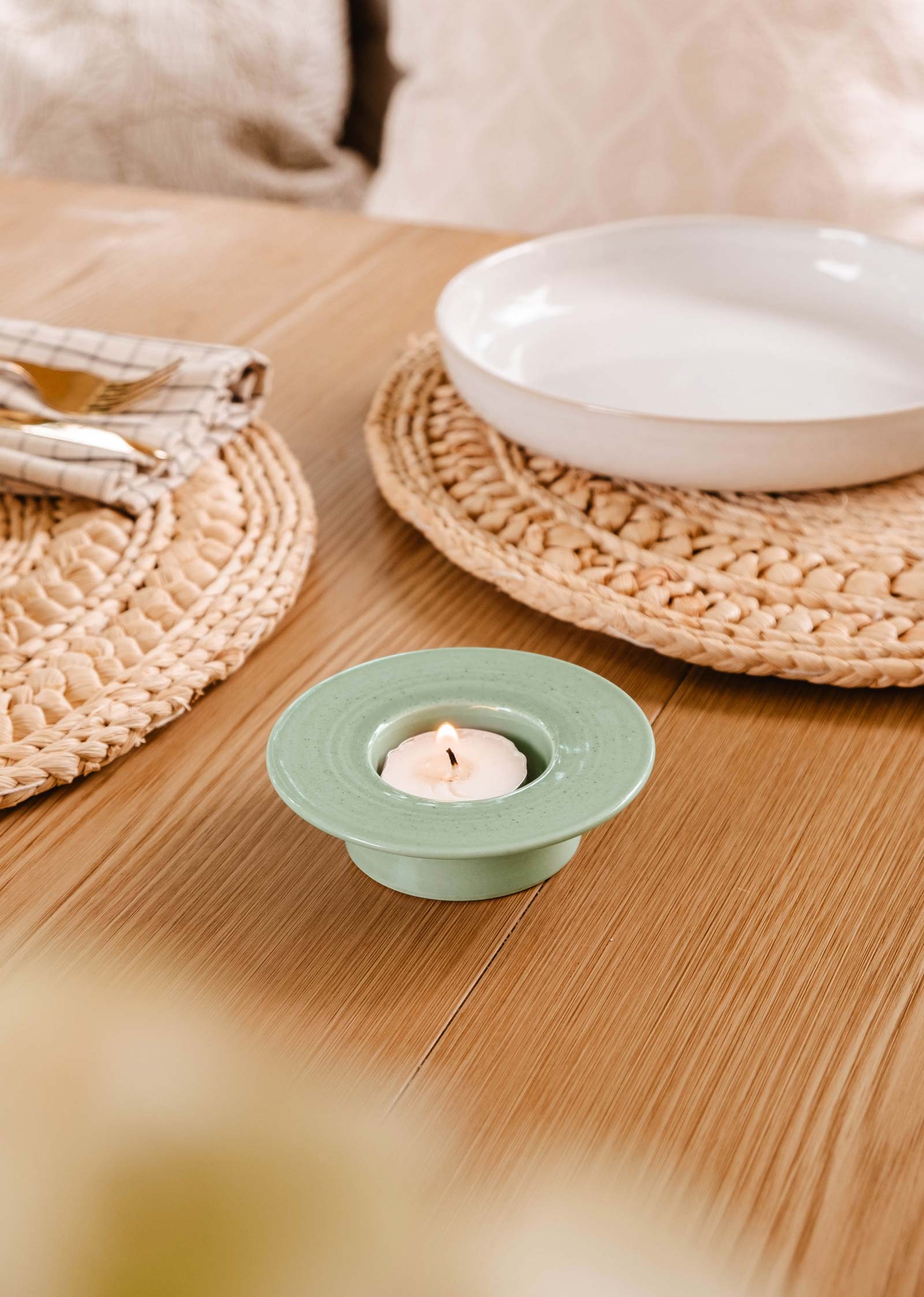 Une petite bougie est allumée dans le Porte-bougies à réchaud rond couleur turquoise de Mimi & August sur une table en bois, entourée d'un décor rustique, de sets de table en osier et d'un plat blanc.