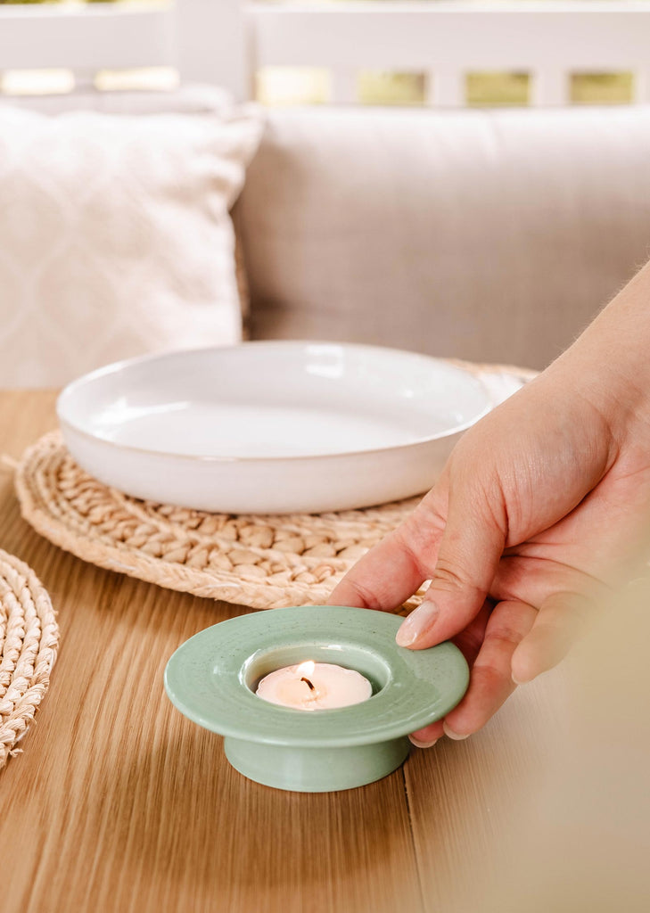 Une main place le porte-bougie à réchaud The Round de Mimi & August avec une bougie allumée sur une table en bois, près d'un napperon tressé et d'un plat blanc, ajoutant au décor rustique.