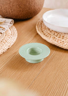 Un petit bol en céramique vert menthe est posé sur une table en bois à côté d'une assiette blanche, du porte-bougies à réchaud rond turquoise de Mimi & August et de sets de table tissés.