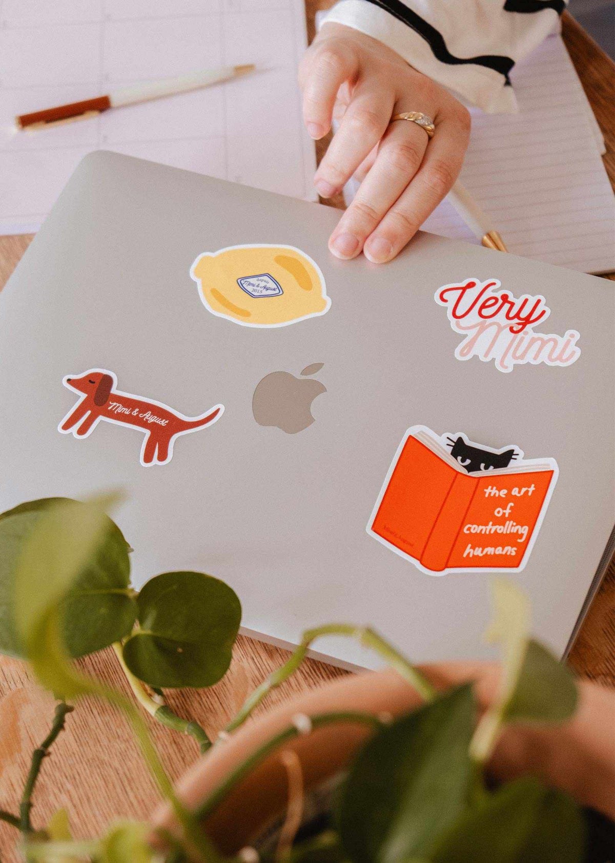 A hand rests on a silver MacBook adorned with colorful stickers, including the Very Mimi - Vinyl Sticker by Mimi & August, next to a plant, notepad, and calendar on a wooden table.