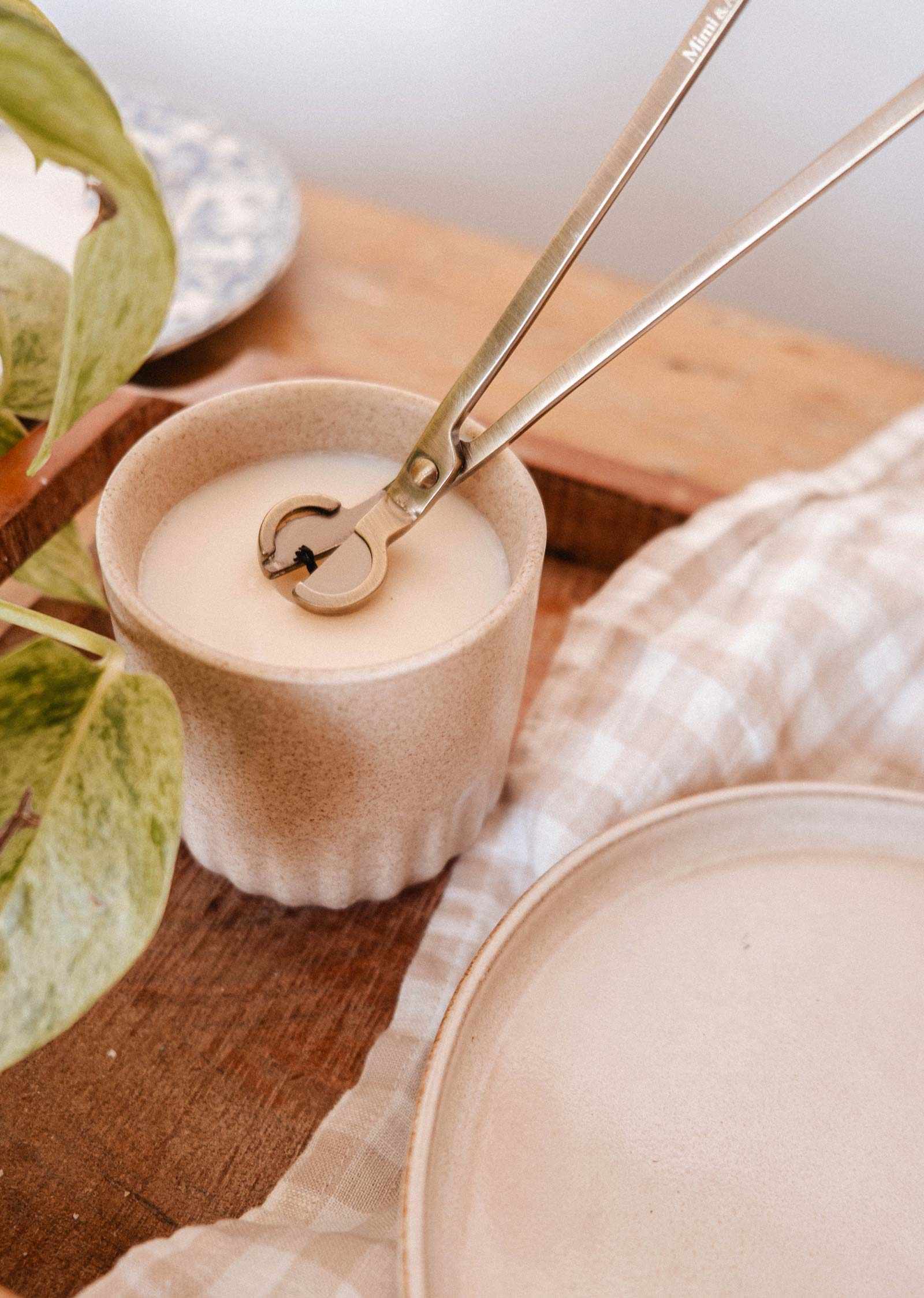 The Mimi & August Wick Trimmer neatly trims candle wicks, shown here with a white candle in a beige ceramic container on a wooden surface—an essential accessory for any cozy space.
