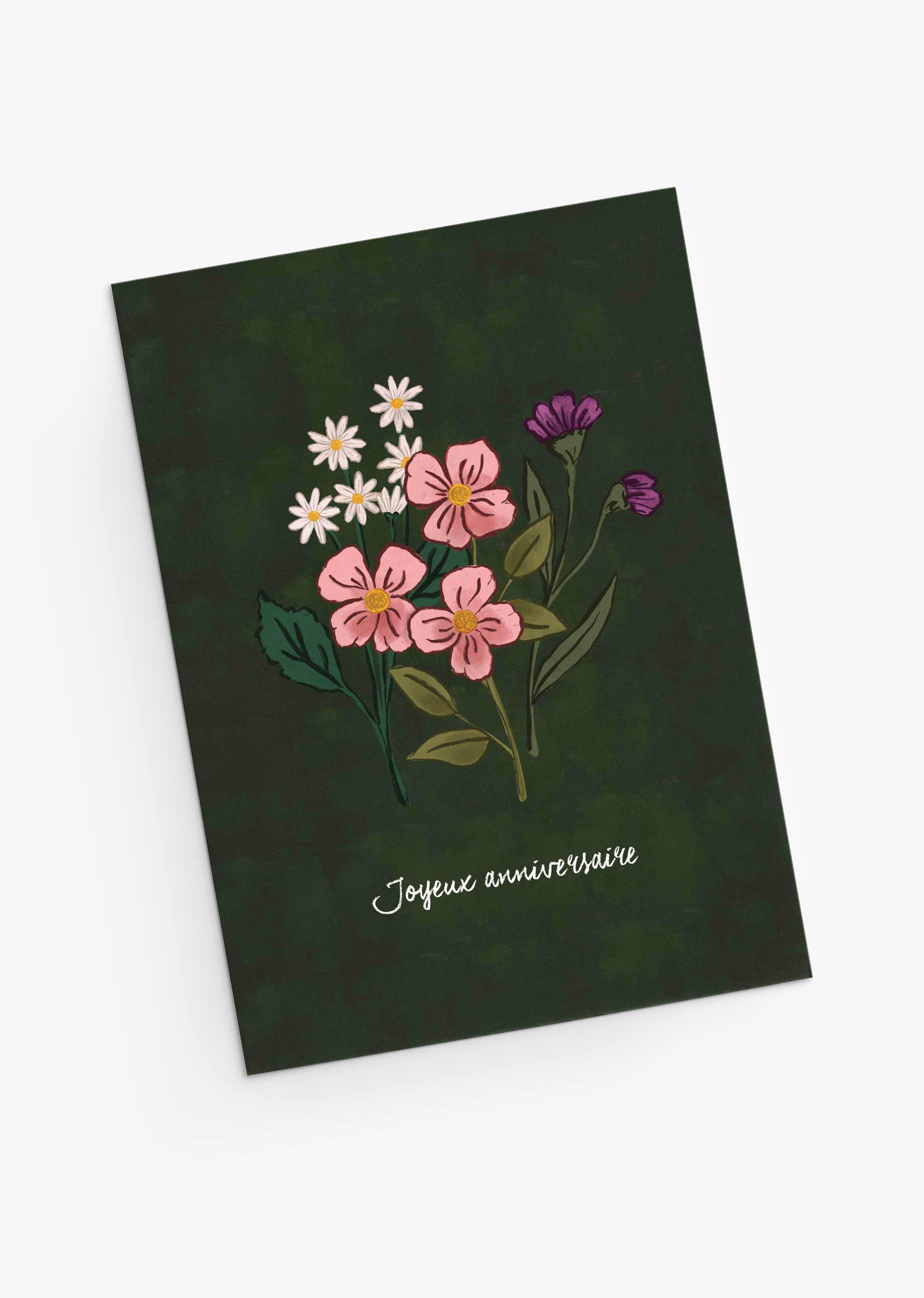 The Wildflower Wishes Greeting Card by Mimi & August is a dark green birthday card with hand-drawn pink, white, and purple blooms and “Joyeux anniversaire” elegantly scripted in white at the bottom.