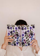A person in a light shirt holds up a spiral-bound notebook with a purple floral cover, concealing their face, against a plain beige background—highlighting the elegant Mimi & August Wildflowers – 2027 Agenda, thoughtfully designed for monthly and weekly planning as your perfect planner for 2027.