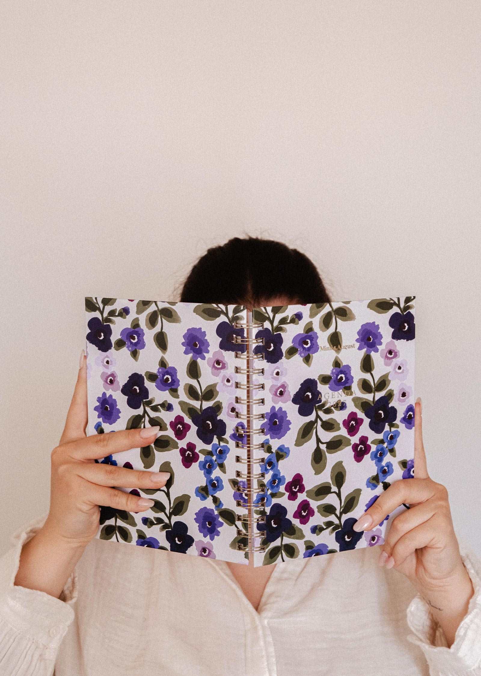 A person in a light shirt holds up a spiral-bound notebook with a purple floral cover, concealing their face, against a plain beige background—highlighting the elegant Mimi & August Wildflowers – 2027 Agenda, thoughtfully designed for monthly and weekly planning as your perfect planner for 2027.