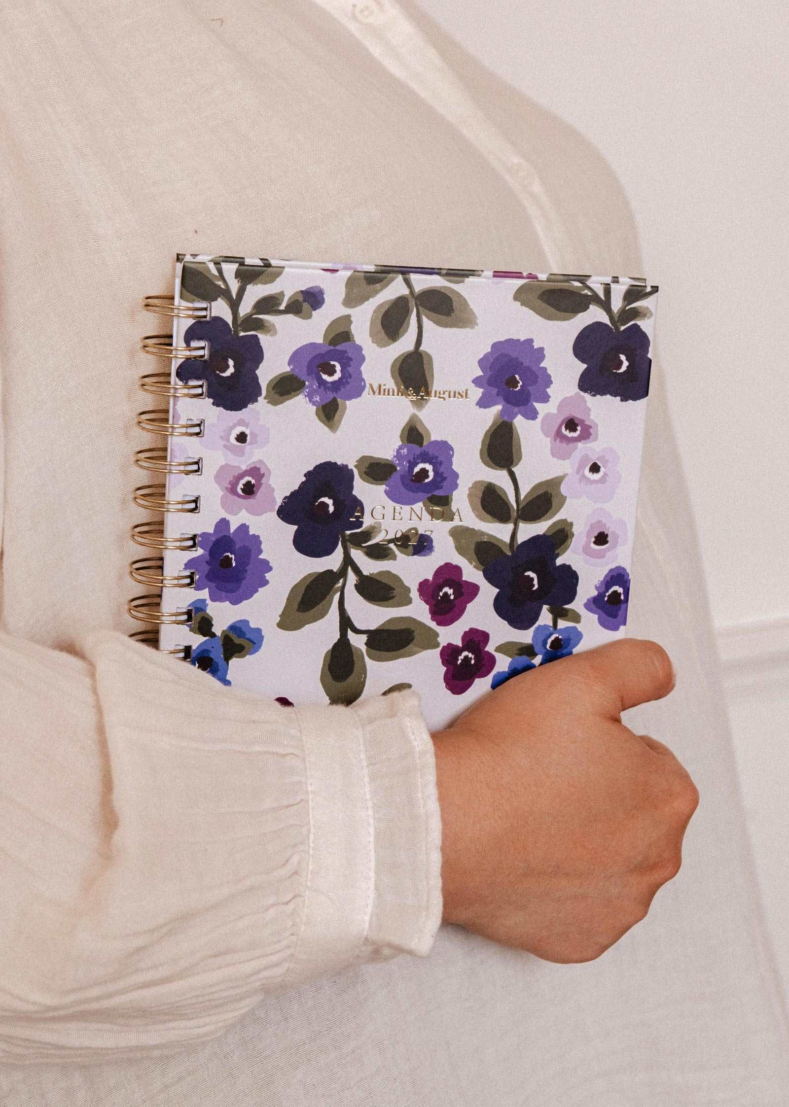 A person in a cream shirt holds the Wildflowers – 2027 Planner by Mimi & August—an illustrated planner featuring purple, blue, and green floral designs—against their chest.