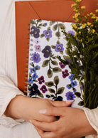 A person holding a brown folder, the Mimi & August Wildflowers – 2027 agenda, a floral spiral notebook, and a bouquet of yellow flowers, dressed in a light-colored long-sleeve shirt—perfect inspiration for anyone looking to organize their monthly and weekly plans or searching for the ideal planner to keep their wildflowers agenda on track.