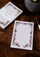 Two Mimi & August Wildflowers floral notepads with a delicate flower border, beside a black pen and a gray flower pot.