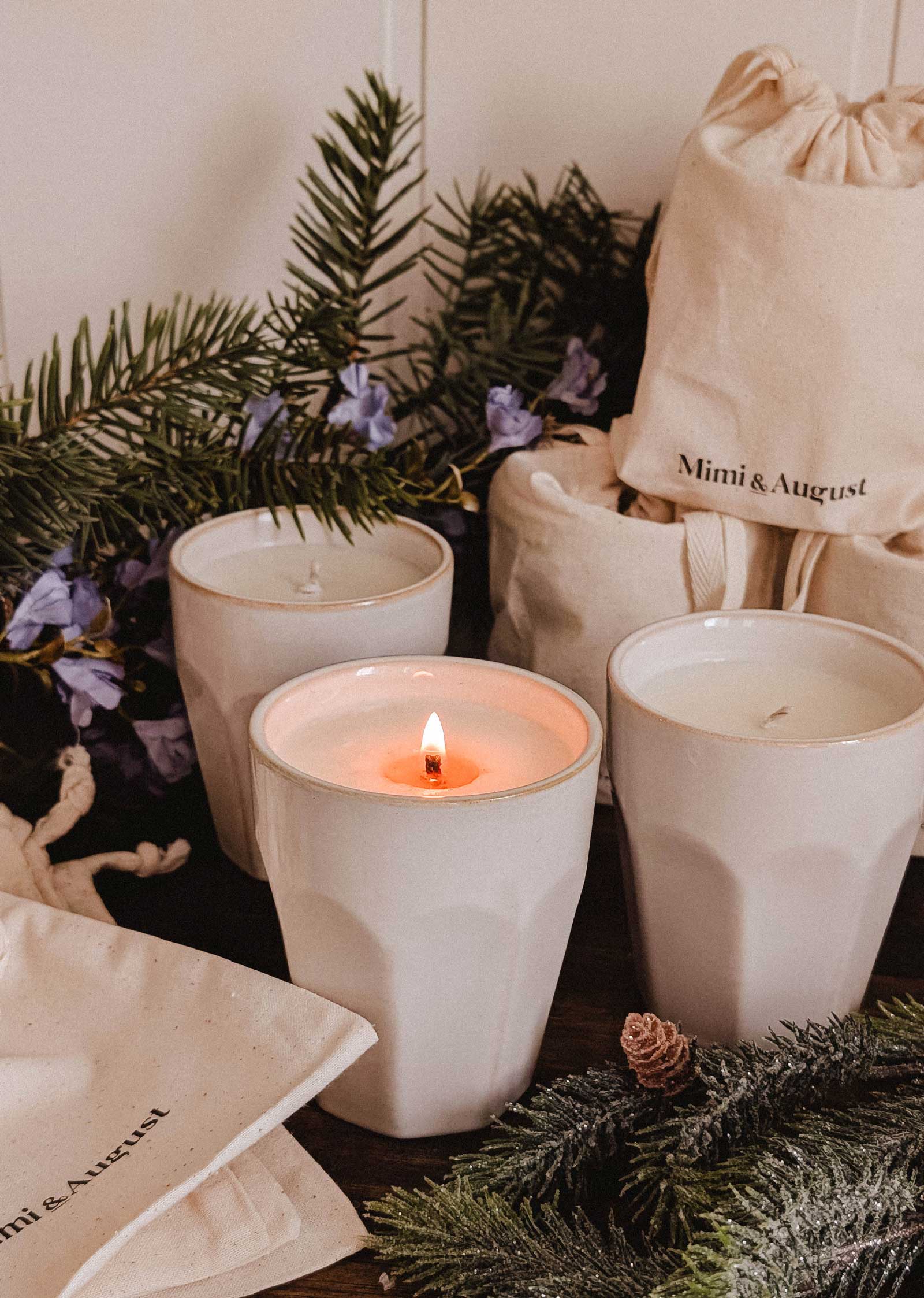 Three Special Edition Candles by Mimi & August—white and in ceramic holders, including a unique scented one (lit), surrounded by pine branches, purple flowers, and branded drawstring bags.