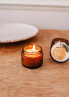 Une bougie de soya Agrumes boisés 2 oz de Mimi & August allumée dans un verre ambré repose sur du bois à côté d'un pot réutilisable ouvert avec étiquette, avec une assiette en céramique blanche en arrière-plan.