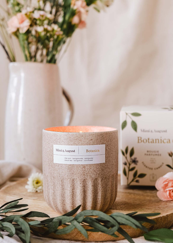 An aromatherapy Mimi & August Botanica - Reusable Candle on a wooden board with a floral arrangement and package in a cozy, homey setting.