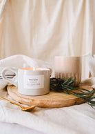 A lit Mimi & August Botanica - Reusable Candle on a wooden tray with green leaves, surrounded by a ceramic mug and soft beige textiles.