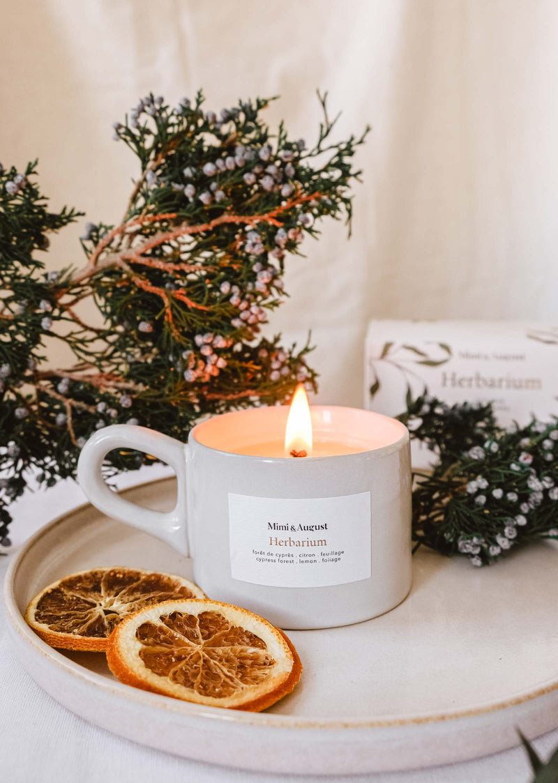 A foliage-filled Mimi & August Herbarium - Reusable Candle exudes a refreshing spring scent as it sits on a plate with orange slices.
