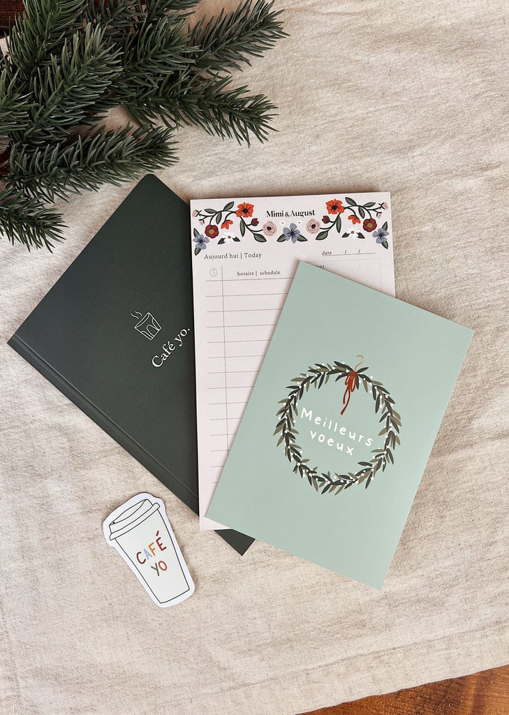 A flat lay of The little elf planner bundle by Mimi & August, a notepad with a floral design, a Meilleurs vœux card with a wreath illustration, a coffee cup sticker, and green pine branches on white fabric.