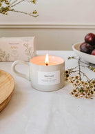 The Artea – Reusable Soy Candle by Mimi & August glows in a mug on a table, surrounded by a box, a bowl of plums, a wooden plate, and sprigs of small white flowers.
