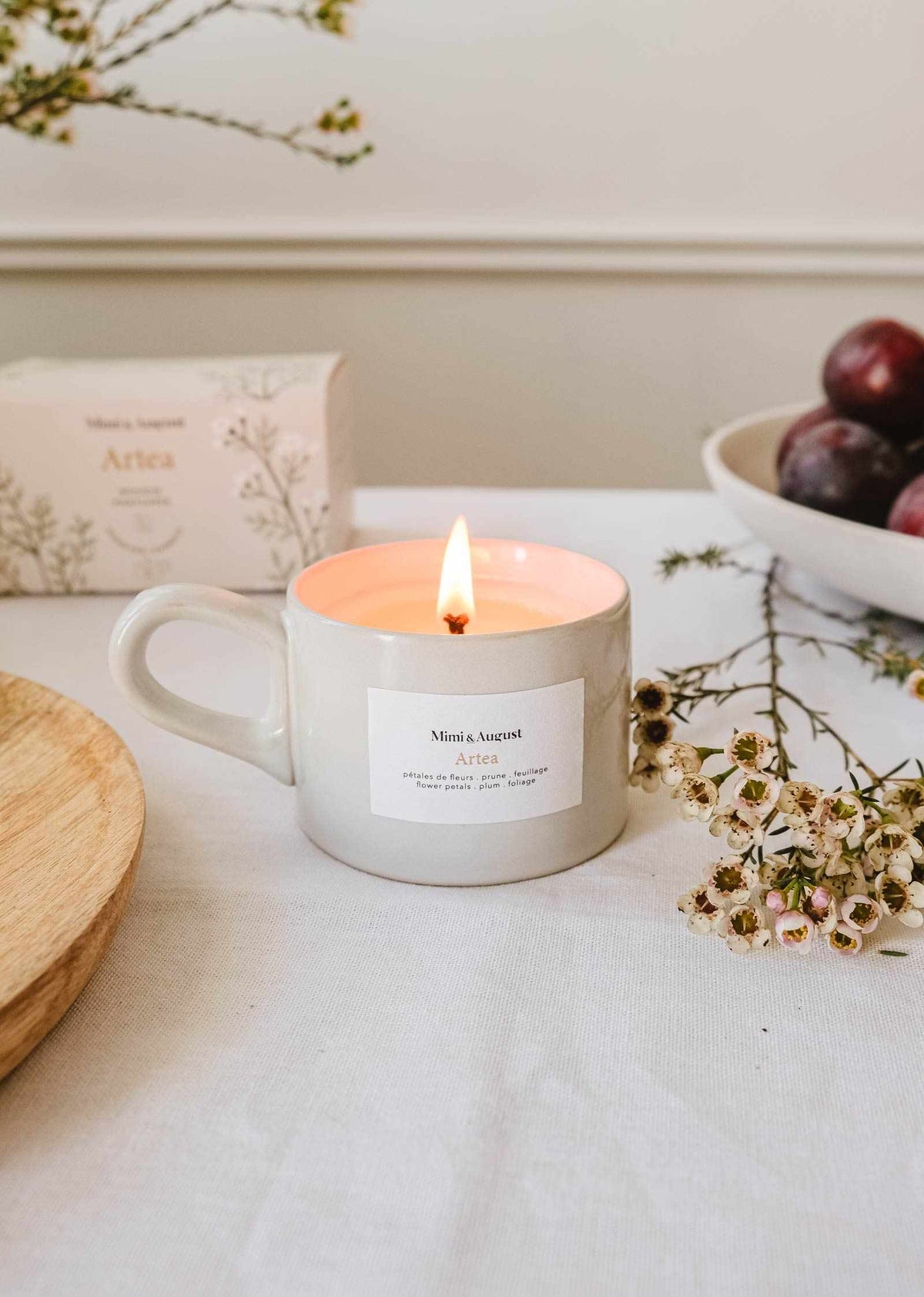 The Artea – Reusable Soy Candle by Mimi & August glows in a mug on a table, surrounded by a box, a bowl of plums, a wooden plate, and sprigs of small white flowers.