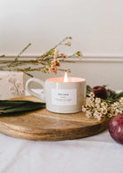 The Artea – Reusable Soy Candle by Mimi & August glows in a ceramic holder atop a wooden tray, surrounded by delicate flowers and leaves, with a plum and a floral box in the background.