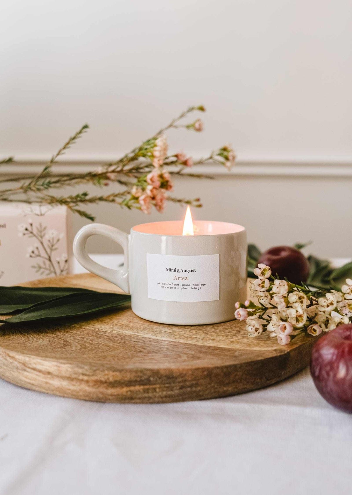 The Artea – Reusable Soy Candle by Mimi & August glows in a ceramic holder atop a wooden tray, surrounded by delicate flowers and leaves, with a plum and a floral box in the background.