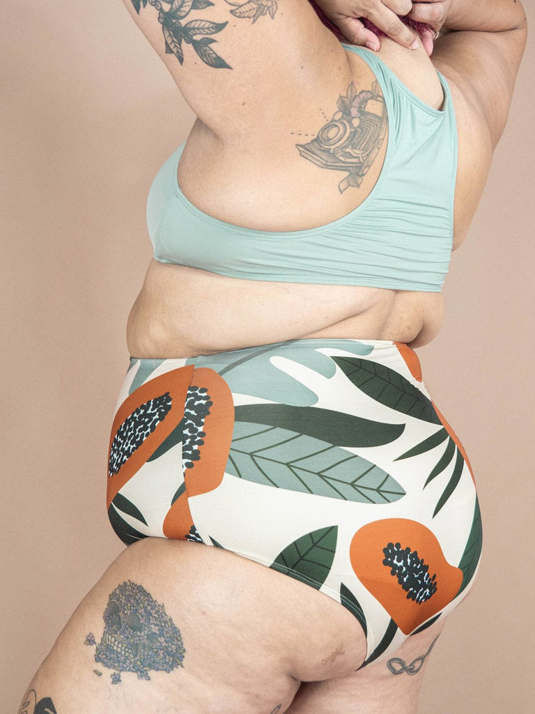 Une femme vêtue d'un bas de bikini Mimi & August Bermudes Papaya Extra High Waist avec des tatouages et un imprimé tropical.