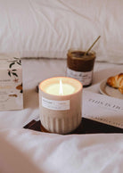 Lit reusable soy coffee candle in ceramic cup on bed with croissant and coffee jar in background