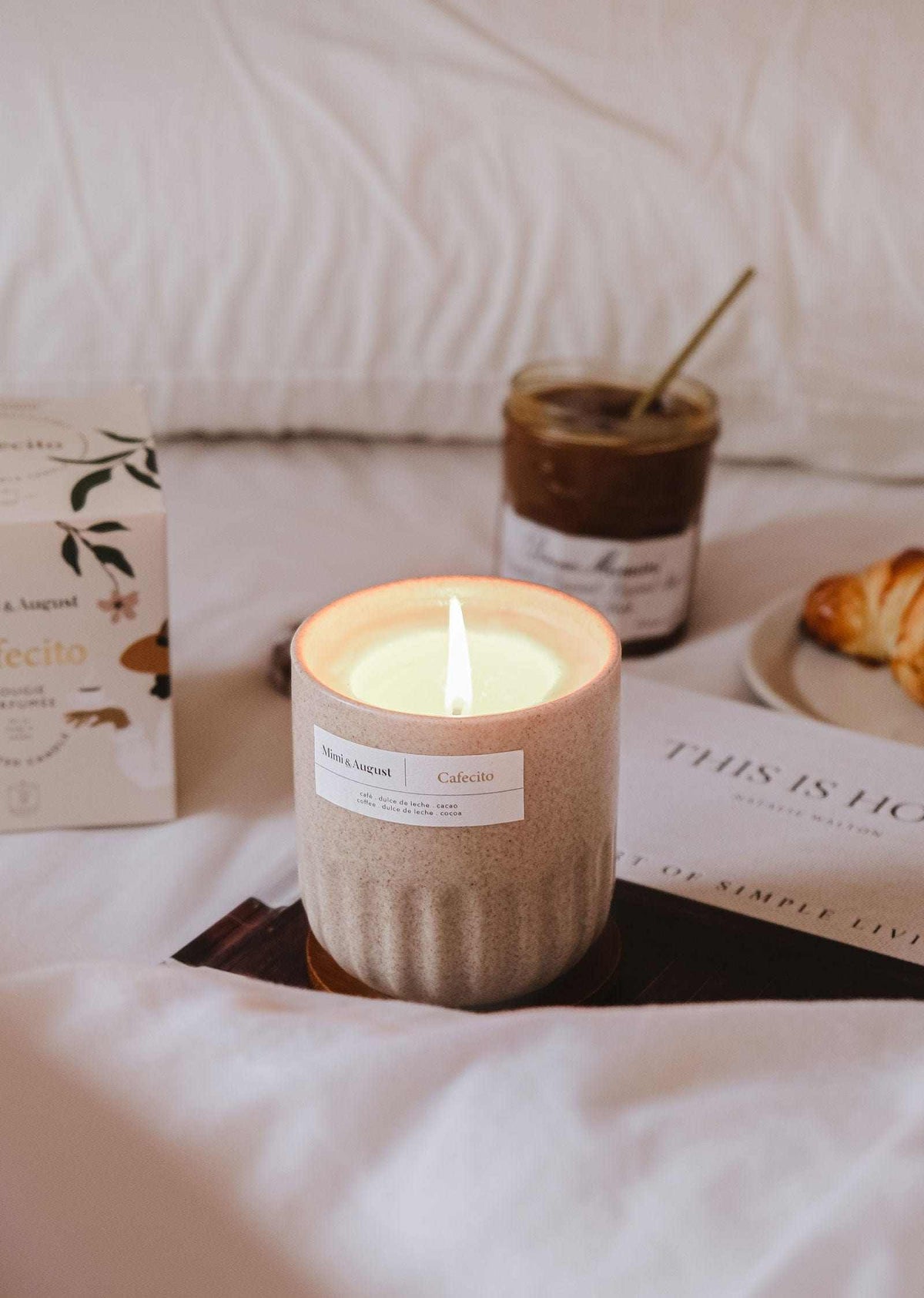Lit reusable soy coffee candle in ceramic cup on bed with croissant and coffee jar in background